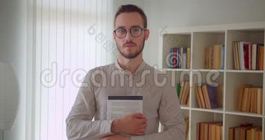 年轻英俊的白种人<strong>男学生</strong>戴着眼镜，举着一本书，看着站在镜头里的镜头。
